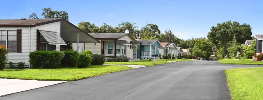 Paved neighborhood street lined with neat park-model homes with carports and green lawns under a blue sky.